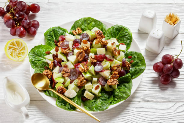 insalata waldorf con uva rossa, sedano, mela verde fresca, noci, uvetta su un cuscinetto di foglie di lattuga fresca su un piatto bianco su un tavolo di legno, cucina americana - healthy eating close up lemon nut foto e immagini stock