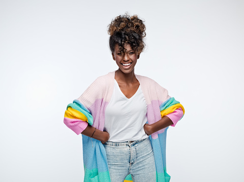Beautiful african young woman wearing rainbow cardigan standing with hands on hip and laughing at camera. Studio portrait on white background.