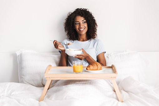 Smiling young african woman having tasty breakfast on a tray in bed