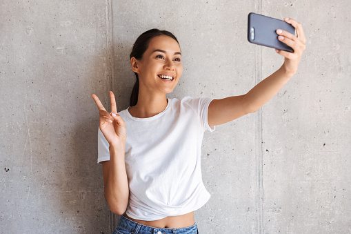Smiling young asian woman taking a selfie with mobile phone over gray wall background