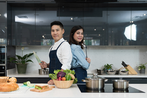 Confident chef and female commis posed to hold a knife with a soup ladle in cooking kitchen and standing back to back each other. Asian couple in apron prepared to cook healthy food on weekends.