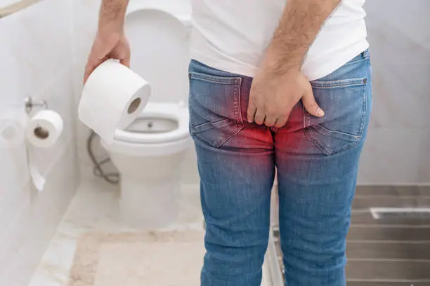 Man suffering from hemorrhoids pain holding toilet paper roll in toilet.