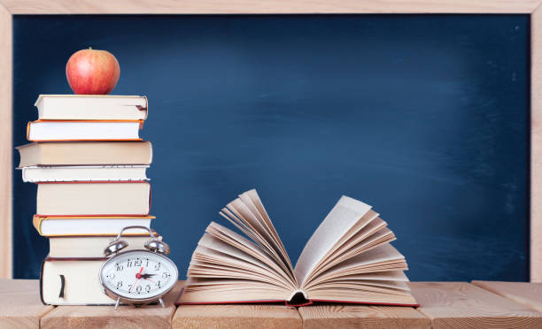 istock Open book with watch, red apple and stack of books on the blackboard background 1327531385