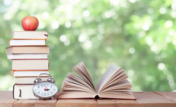 Photo of Open book with watch, red apple and stack of books on the nature background