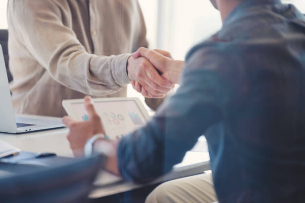 Close up of Businessmen shaking hands. Close up of Businessmen shaking hands. Both are dressed in casual clothing and one is holding a digital tablet with graphs and charts. Shot through a window. There is also a laptop computer on the table business consultation stock pictures, royalty-free photos & images