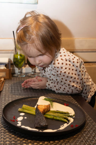 A little girl in a restaurant looks at a plate of baked salmon and asparagus, decorated with sauce and caviar of flying fish, on the table in front of her, she looks very carefully at how the fish dish is decorated, and wants to try it faster A little girl in a restaurant looks at what the official brought on a plate with baked salmon and asparagus, decorated with sauce and caviar of flying fish, on the table in front of her she looks very carefully at how the fish dish is decorated Her daughter looks at the food with hungry eyes and wants to try it as quickly as possible eating asparagus stock pictures, royalty-free photos & images