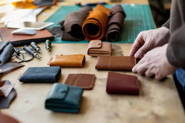 Craftsman hands laying out exclusive leatherwork on wooden table at leather workshop. Male tanner preparing handwork stuff accessories to sending clients or making promotional photo for advertising