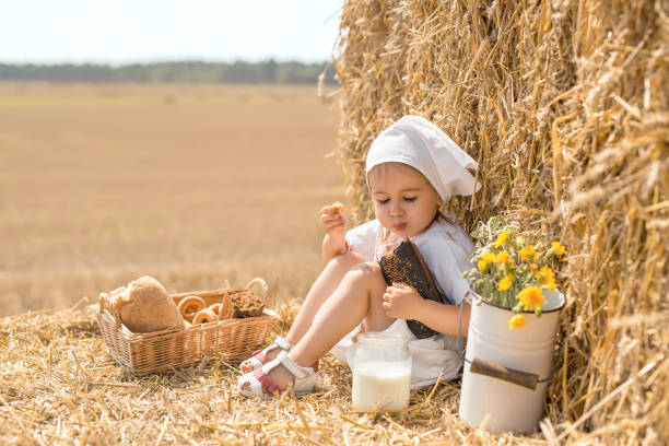 a girl in a white scarf is sitting in a field on a haystack and eating bread - breadcrumb navigation imagens e fotografias de stock