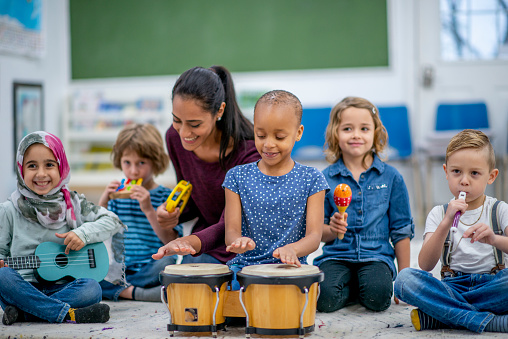 A diverse group of children sits on the carpet and play musical instruments. They are smiling, having a good time.