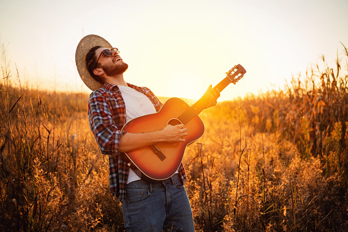 One young handsome Caucasian man playing guitar while standing on the field during a sunset.