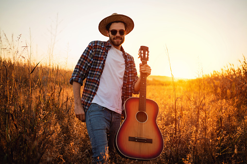 One young handsome Caucasian man with guitar walking on the field during a sunset.
