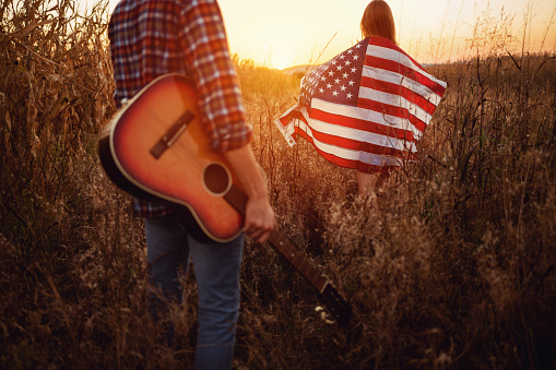 Two people, beautiful young couple, with American flag and guitar walking on the field during a sunset.