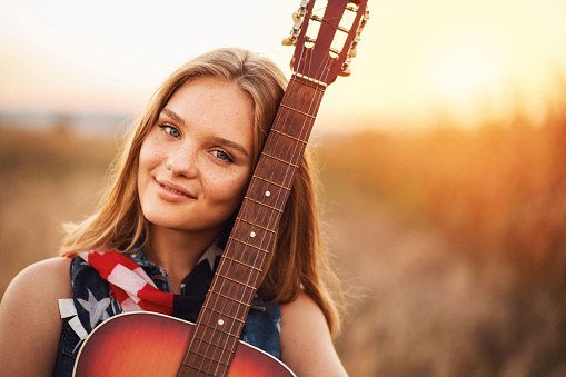 One young beautiful American woman with a guitar standing on the field at the sunset and looking at camera.