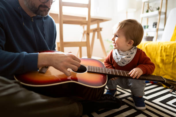 Unrecognizable father playing guitar with his little son at home Caucasian father playing  acoustic guitar with his little son while sitting on the floor at home. father and son guitar stock pictures, royalty-free photos & images
