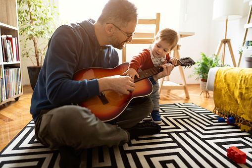 Caucasian father playing  acoustic guitar with his little son while sitting on the floor at home.