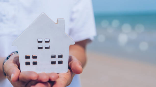 istock Hand holding wooden home model on beach background 1327565332