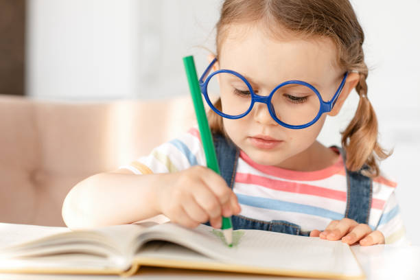 istock Smart child girl with glasses draws colored pencils at table in white kitchen. Developing children's skills, kid hobbies, education and learning, back to school. Background, copy space 1327578691