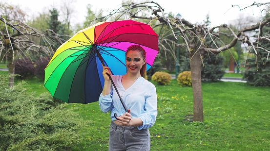 Girl in a bluish shirt with bright makeup and long colored braids. Holding an umbrella in the colors of the rainbow on a flowered park enjoying the coming spring