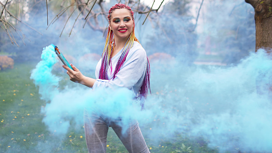 A cheerful girl in a shirt and jeans with bright rainbow braids and unusual makeup. She dances hiding in thick blue artificial smoke against the backdrop of spring park