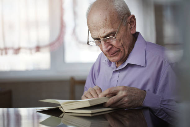 istock Hoary pensioner wearing glasses sitting at table and reading book 1327586837