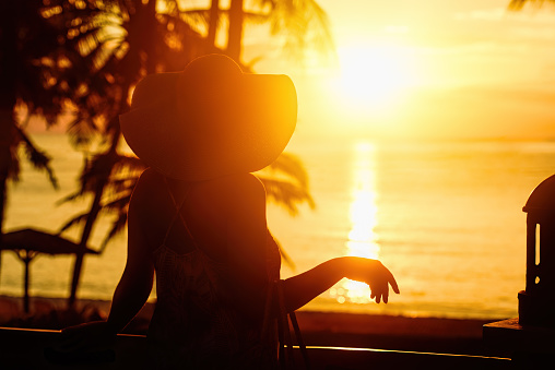 Vacation Summer Holiday Concept. Woman in hat watching the beautiful scenic sunset on the terrace luxury hotel.