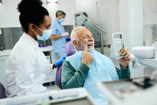 Mature man using mirror and looking at his teeth after dental procedure at dental clinic.