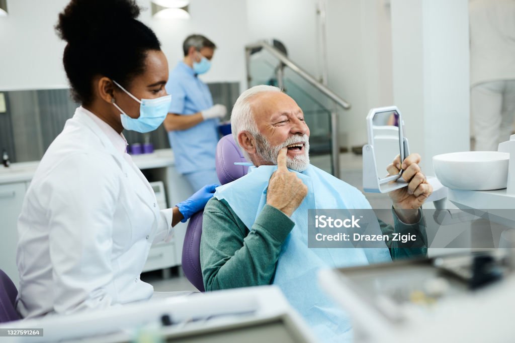 Senior man looking his teeth in a mirror after dental procedure at dentist's office. Mature man using mirror and looking at his teeth after dental procedure at dental clinic. Dentist's Office Stock Photo
