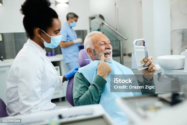 Senior Man Looking His Teeth In A Mirror After Dental Procedure At Dentists Office Stock Photo - Download Image Now