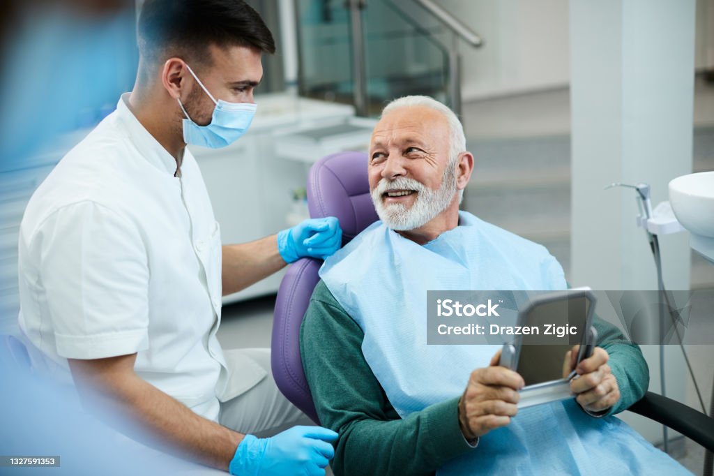 Satisfied senior man communicating with his dentist after dental procedure at dentist's office. Happy senior man talking to his dentist while being satisfied with dental procedure at dentist's office. Dentist Stock Photo