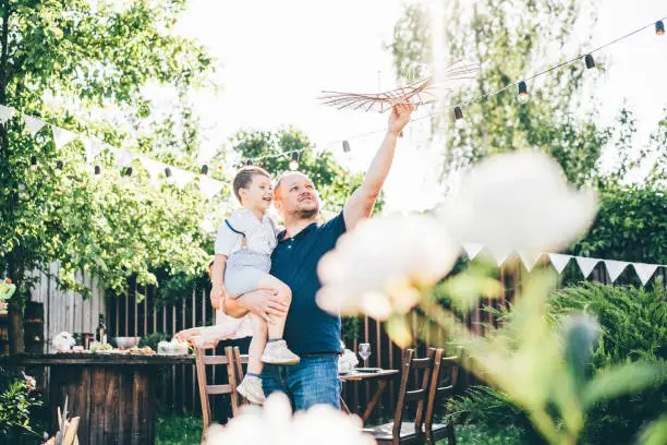 smiling father in t-shirt plays with toy plane holding adorable little son in arms in summerhouse green backyard decorated with garlands on sunny day