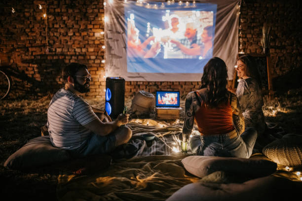 Movie night Three people, man and two women sitting together on the ground in back yard outdoors. They are watching a movie together. body adornment rear view young men men stock pictures, royalty-free photos & images