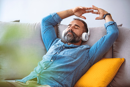 Shot of a mature man wearing headphones while relaxing at home. I'm in my happy place right now. Happy mid adult man using smartphone device while sitting on sofa at home