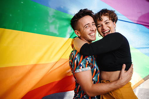 Smiling queer couple standing against a rainbow pride flag. Young LGBTQ couple embracing each other while standing together. Two non-conforming lovers celebrating gay pride together.