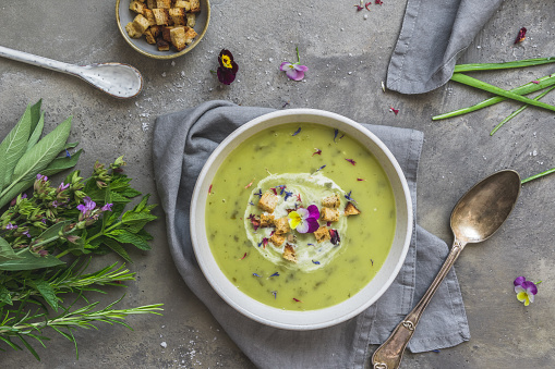 Creamy herbes soup with sorrel, edible blooms and croutons as topping. Gray concrete background. Top view.