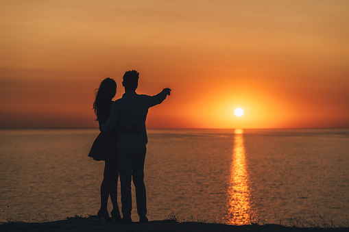 The young couple standing on the sea shore on the picturesque sunset background