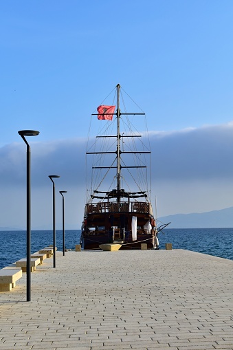 Wooden sailboat with lowered sails under Albanian flag at pier on background of blue sea and sky