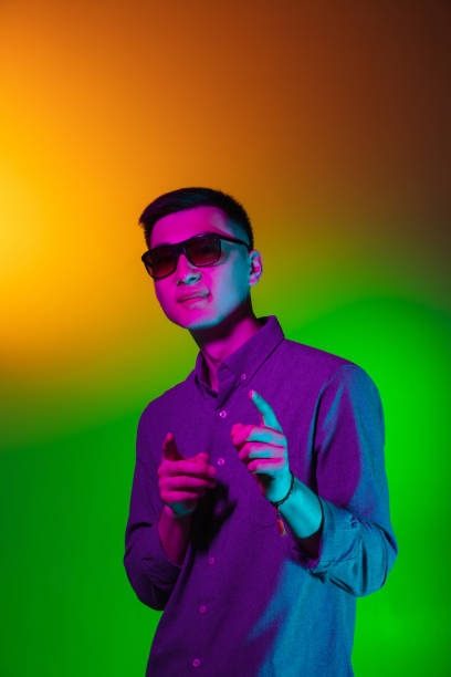 istock Handsome young man in sun glasses isolated on studio background in gradient green yellow neon light, colour filter. Concept of human emotions, facial expression. 1327622138