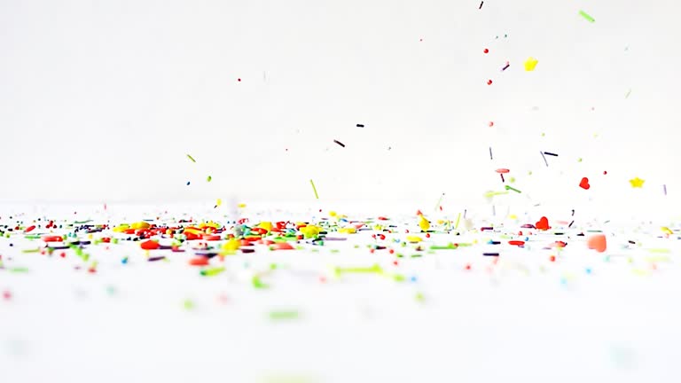 Multi-colored differently shaped sweet sprinkles are poured on a wooden white table on a white background. Slow motion bright fun video.