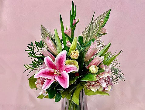 Horizontal centre placed still life of pink and green flower bouquet in vase with lillies against light pink wall