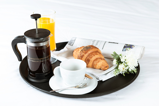 Breakfast with croissant, french press coffee, orange juice, flowers and newspaper on bed with white sheets.