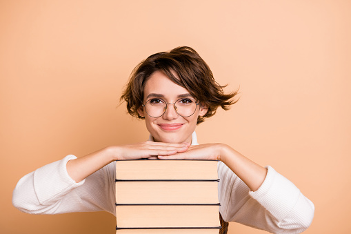 Portrait of young beautiful happy positive cheerful smiling girl in glasses with pile on books isolated on beige color background.