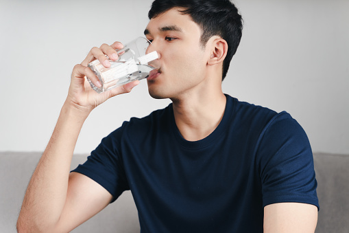 handsome asian man drinking a glass of water on the sofa at living room.