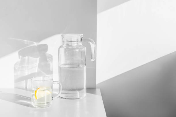 agua fresca con rodajas de limón en garrafa y vaso sobre mesa blanca con sombras y luz. - botella de boca ancha fotografías e imágenes de stock