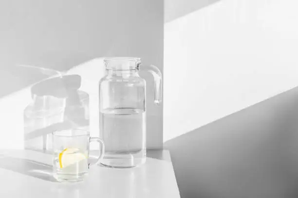 Fresh water with lemon slices in carafe and glass on white table with shadows and light.