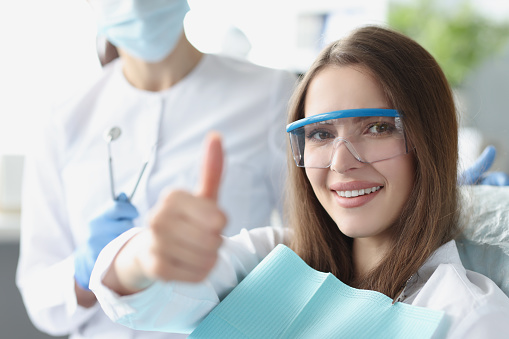 Young woman in safety glasses sitting in dental chair and showing thumbs up. Quality dental care concept