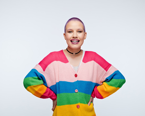 Cheerful young woman wearing rainbow cardigan standing with hands on hips and laughing at camera. Studio portrait on white background.