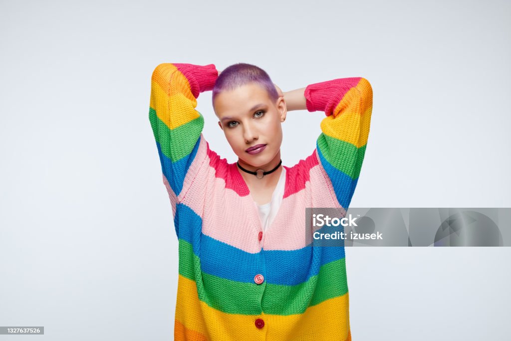 Friendly young woman with short purple hair and rainbow cardigan Happy young woman wearing rainbow cardigan, standing with raised arms and smiling at camera. Studio portrait on white background. Transgender Person Stock Photo