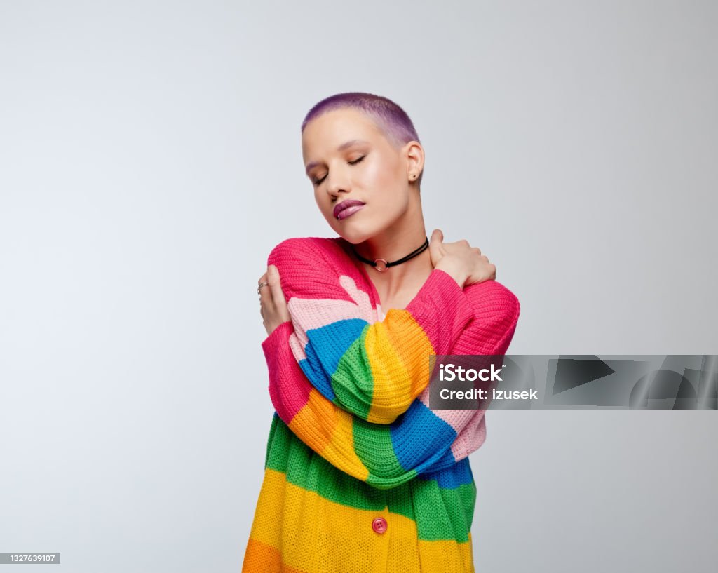 Short hair woman with rainbow sweater Young woman wearing rainbow cardigan hugging self. Studio portrait on white background. Transgender Person Stock Photo