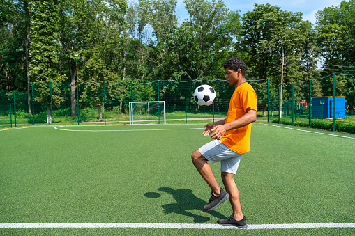 Young Brazilian football player on sport field kicking and stuffing soccer ball on workout outdoors in summer morning, side view