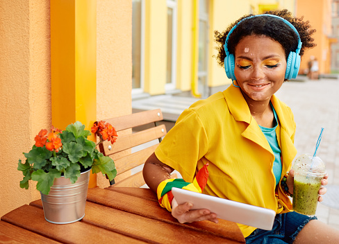 Positive young woman with vitiligo wearing a blue headphones and trendy clothes sits in outdoor summer cafe and drinks a vitamin cocktail looking into her tablet